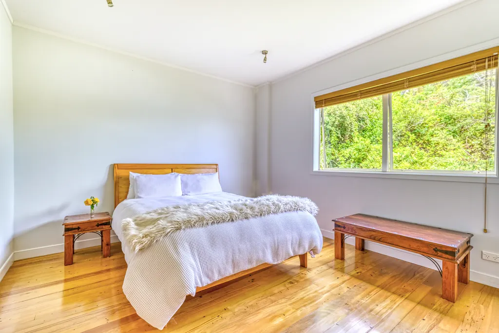 Bright bedroom with a timber bed, native bush outside the window and honey-toned floorboards